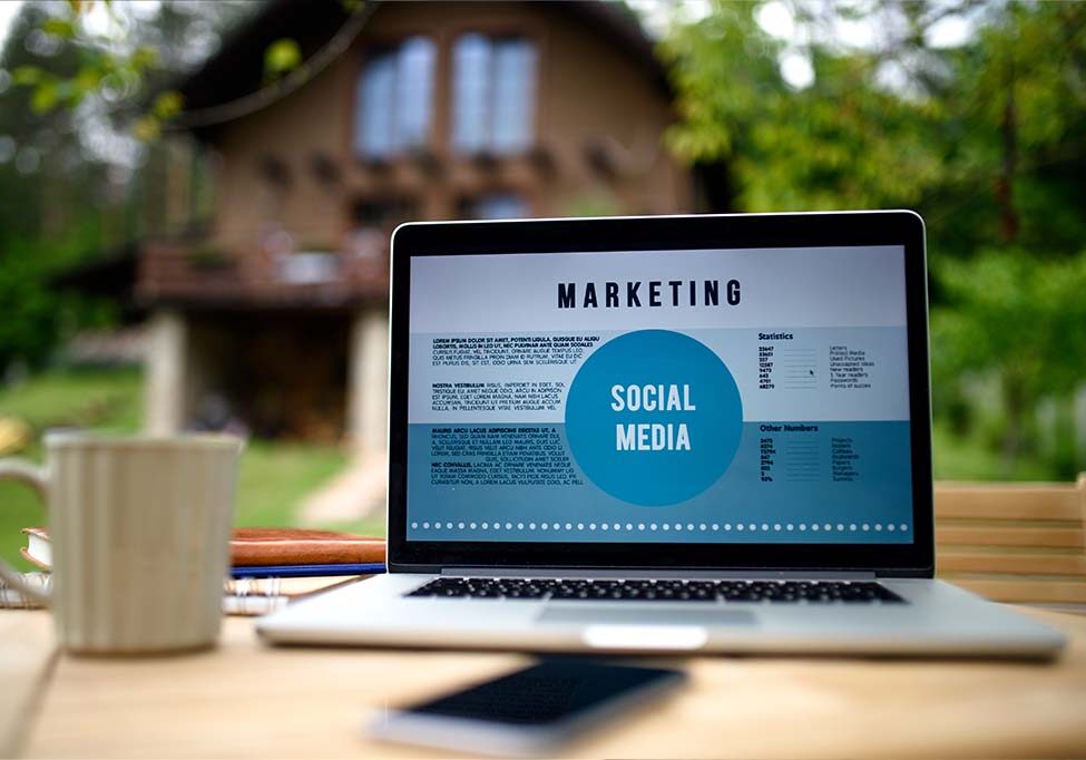 Social Media Marketing_01 Social Media Marketing mit der Videoproduktion Stuttgart. Das Bild zeigt einen Laptop, der auf einem Tisch im Freien steht. Auf dem Bildschirm ist eine Präsentation oder Grafik mit dem Titel „Marketing“ zu sehen, die das Thema „Social Media“ in den Mittelpunkt stellt. Der Text und die Gestaltung deuten darauf hin, dass es sich um Inhalte handelt, die die Bedeutung oder Strategien des Social-Media-Marketings darstellen. Im Hintergrund ist ein Haus mit einem Garten erkennbar, das eine entspannte und natürliche Atmosphäre vermittelt. Auf dem Tisch befinden sich weitere Gegenstände wie eine Tasse und ein Smartphone, was den Eindruck eines Arbeitsplatzes im Freien verstärkt. Die Kombination aus Technik, Natur und Arbeitsmaterialien suggeriert eine moderne, flexible Arbeitsweise, bei der Marketing- oder Social-Media-Strategien auch außerhalb eines Büros bearbeitet werden können. Das Bild symbolisiert Effizienz und Kreativität in einer inspirierenden Umgebung.